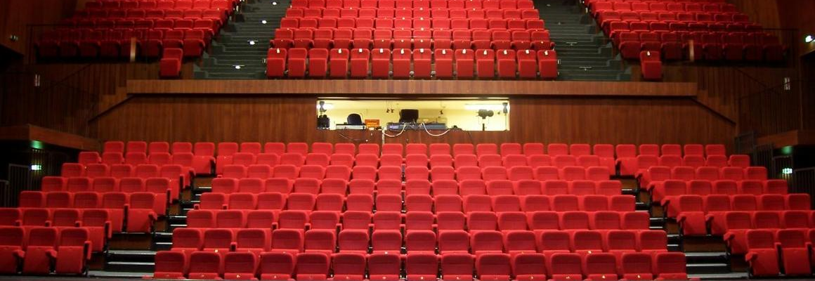 Salle de spectacle avec sièges rouges, scène et rangées de gradins.