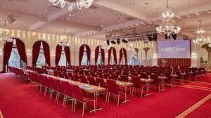 Salle de conférence avec rangées de chaises, tables, écran, lustres et rideaux.