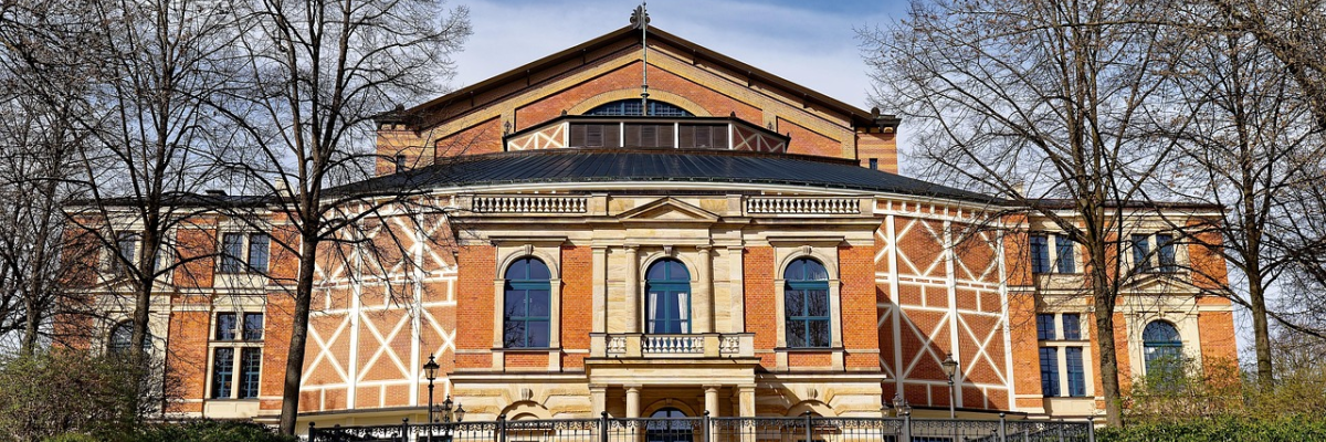 Bâtiment en brique avec façade blanche, toit foncé, entouré d'arbres et de fleurs, avec une entrée centrale.