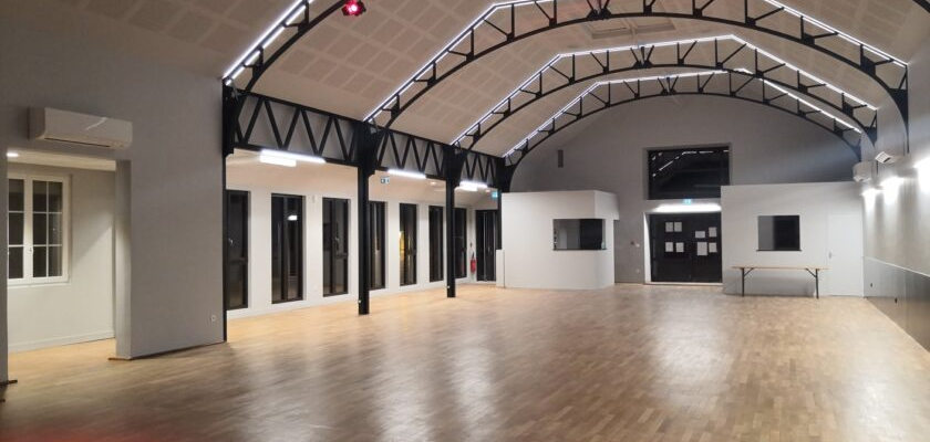 Grande salle avec plancher en bois, piliers noirs, arches métalliques, projecteurs et portes vitrées.