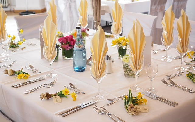 Table de banquet avec vaisselle, verres, serviettes en forme de feuilles, bouteilles, fleurs et chaises.