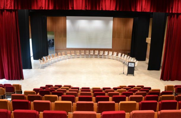 Salle de conférence avec rangées de chaises, écran, podium et rideaux rouges.