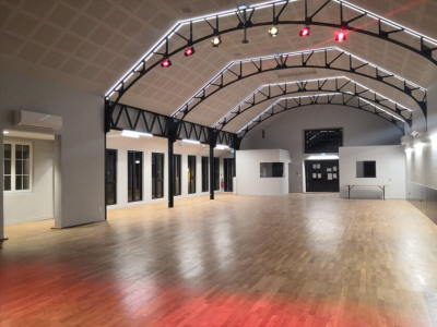 Grande salle avec plancher en bois, piliers noirs, arches métalliques, projecteurs et portes vitrées.
