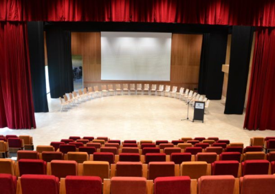 Salle de conférence avec rangées de chaises, écran, podium et rideaux rouges.